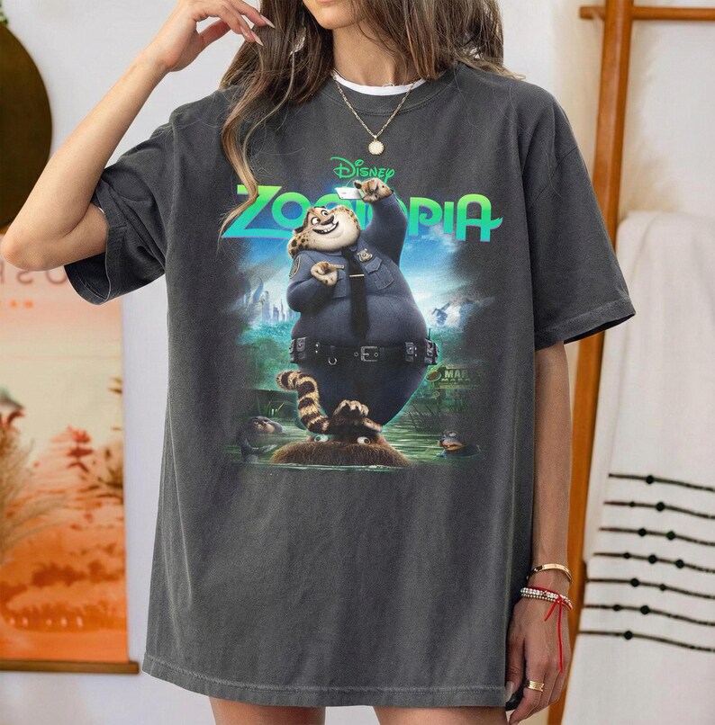 K&ouml;nnte beinhalten: Ein anthrazitfarbenes Kurzarm-T-Shirt mit einem Grafikdruck der Figur Officer Clawhauser aus dem Film Zoomania. Das Design zeigt das gr&uuml;ne Zoomania-Logo &uuml;ber der Figur, die eine blaue Polizeiuniform tr&auml;gt.