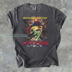 May include: A charcoal gray short-sleeve t-shirt featuring a graphic print of Spider-Man fighting a green creature. The text BRAND NEW DAY appears in yellow above the action scene, and the red and blue SPIDER-MAN logo is printed at the bottom.