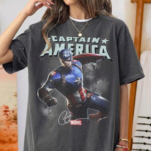 Puede incluir: Camiseta gris oscuro con un dise&ntilde;o de Capit&aacute;n Am&eacute;rica en acci&oacute;n. El texto "CAPTAIN AMERICA" est&aacute; impreso encima de la imagen, con el logo de Marvel y una firma debajo. La camiseta tiene un aspecto vintage.