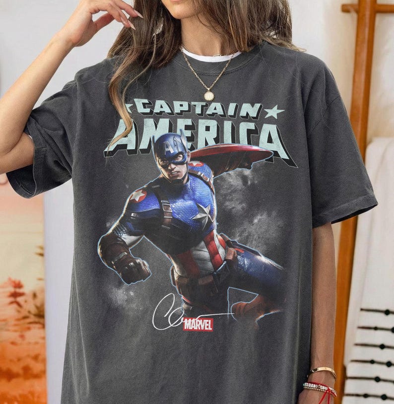 Puede incluir: Camiseta gris oscuro con un dise&ntilde;o de Capit&aacute;n Am&eacute;rica en acci&oacute;n. El texto "CAPTAIN AMERICA" est&aacute; impreso encima de la imagen, con el logo de Marvel y una firma debajo. La camiseta tiene un aspecto vintage.