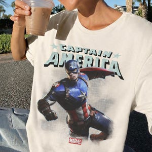 Puede incluir: Camiseta color crema con un dise&ntilde;o de Capit&aacute;n Am&eacute;rica en acci&oacute;n y el texto "CAPTAIN AMERICA" en la parte superior. El logotipo de Marvel est&aacute; en la parte inferior. La camiseta es de un material suave.
