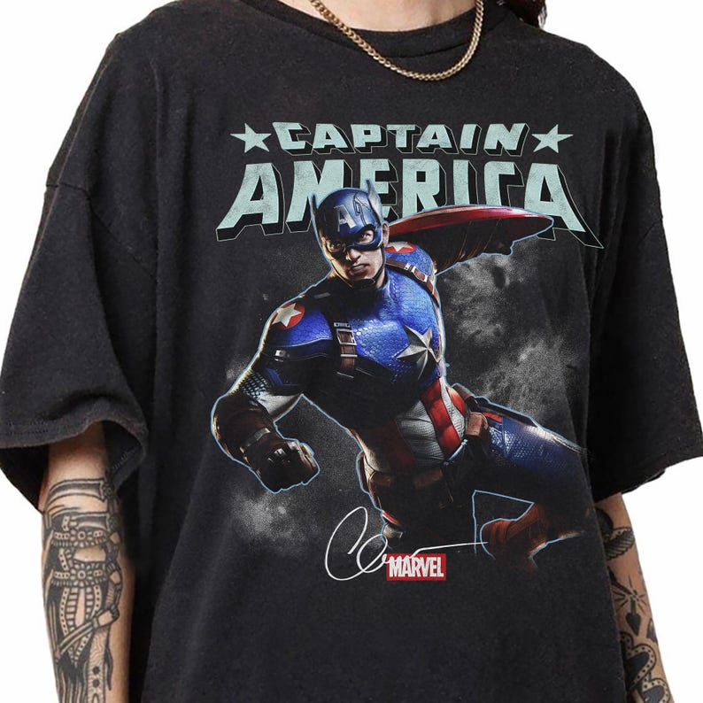 Puede incluir: Camiseta negra con Captain America en una pose din&aacute;mica, con su escudo. El dise&ntilde;o incluye el texto "CAPTAIN AMERICA" en una fuente audaz y estilizada, junto con el logotipo de Marvel y una firma.