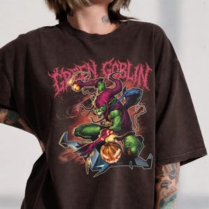 May include: Dark brown t-shirt featuring a Green Goblin graphic. The design includes the text "GREEN GOBLIN" in a pink, stylized font, and an illustration of the Green Goblin character with a pumpkin. The shirt has a vintage, worn look.