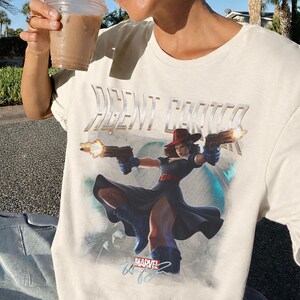 Pu&ograve; includere: T-shirt color crema con una grafica di Agent Carter della Marvel, che tiene due pistole con fiamme. Il testo "AGENT CARTER" &egrave; sopra l'immagine. Il logo Marvel &egrave; in basso.