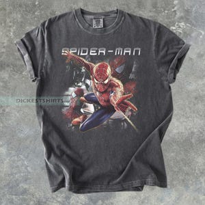 May include: A charcoal gray short-sleeve t-shirt featuring a graphic print of the character Spider-Man in a dynamic pose. The text SPIDER-MAN is printed in white above the character, and the text DICESTSHIRTS is visible on the left side.