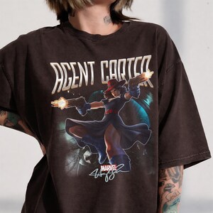 Pu&ograve; includere: T-shirt marrone scuro con una grafica di Agent Carter della Marvel, che tiene due pistole con fiamme. Il design include il testo "AGENT CARTER" in bianco e il logo Marvel.