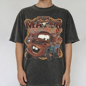 May include: Dark gray t-shirt featuring a graphic of Tow Mater from the movie Cars. The design includes the text "TOW MATER" in a western-style font, with a rope border and a bull skull. The shirt has short sleeves.