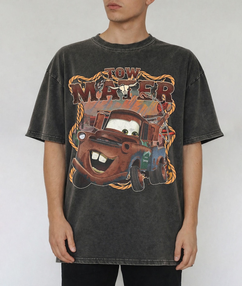 May include: Dark gray t-shirt featuring a graphic of Tow Mater from the movie Cars. The design includes the text "TOW MATER" in a western-style font, with a rope border and a bull skull. The shirt has short sleeves.