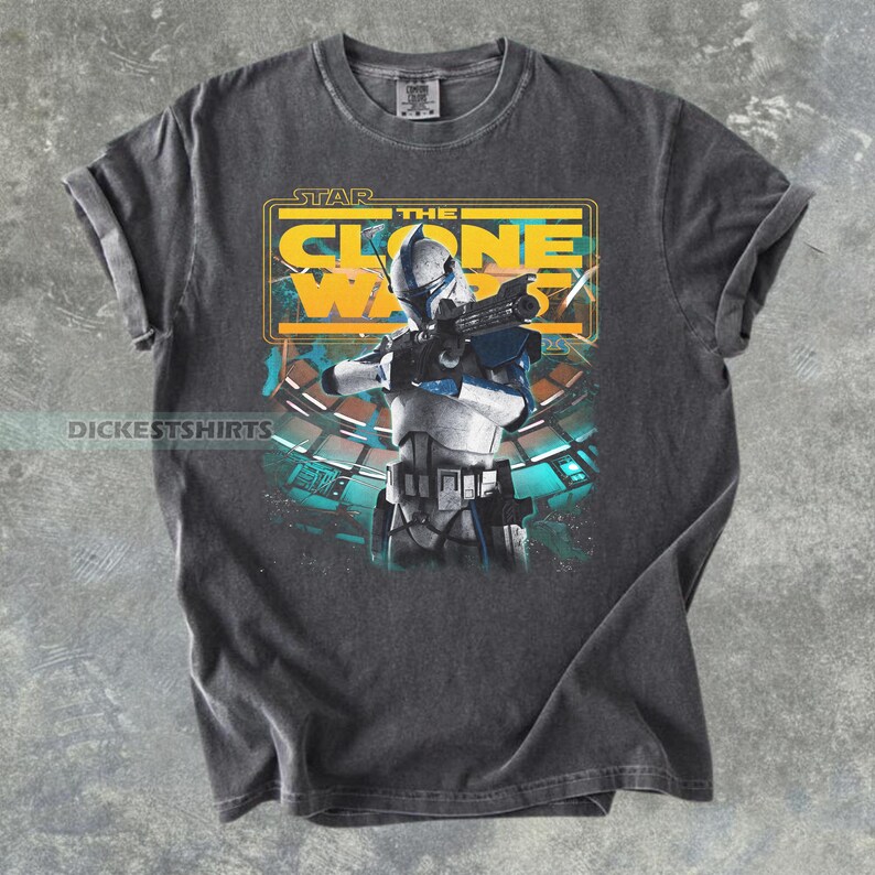 May include: A charcoal gray t-shirt featuring a graphic print of a white and blue clone trooper holding a blaster. The text STAR THE CLONE WARS is printed in yellow above the character. The design includes a teal and black background element.