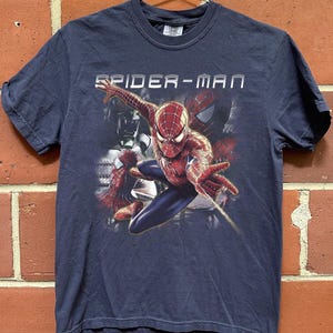 May include: A dark blue short-sleeve t-shirt featuring a graphic print of the character Spider-Man in a dynamic pose with a web-shooting hand gesture. The text SPIDER-MAN is printed in white above the character, with additional shadowy figures visible.