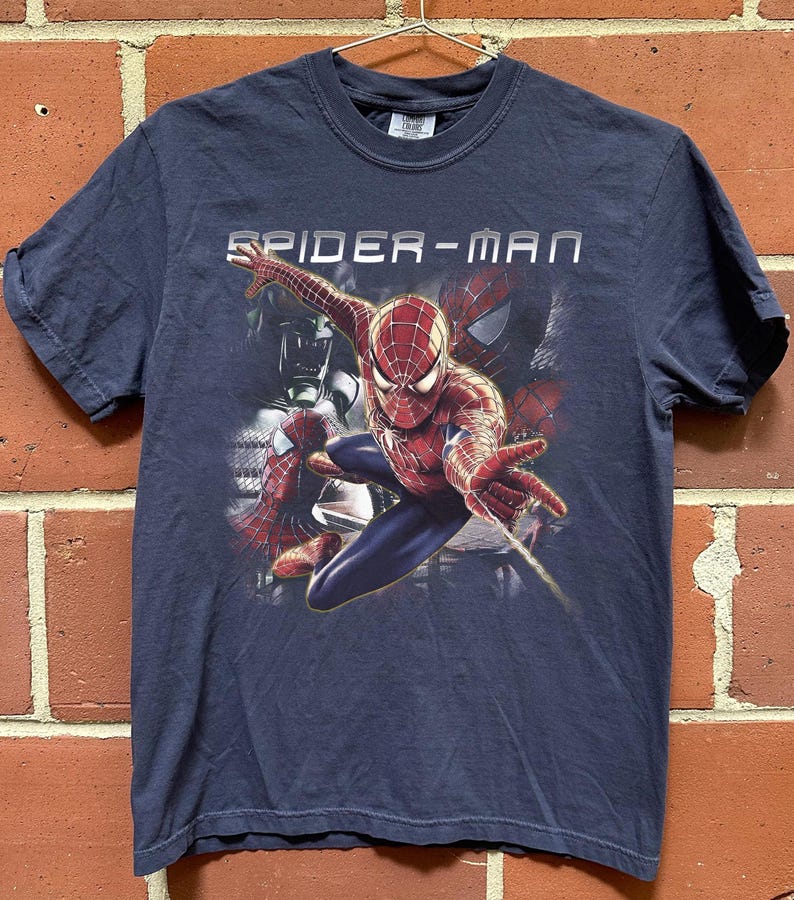 May include: A dark blue short-sleeve t-shirt featuring a graphic print of the character Spider-Man in a dynamic pose with a web-shooting hand gesture. The text SPIDER-MAN is printed in white above the character, with additional shadowy figures visible.