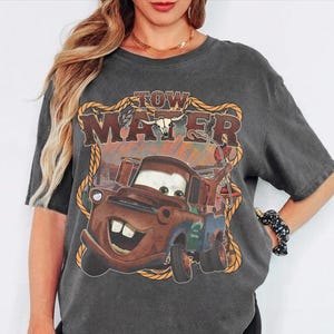 May include: Dark gray t-shirt featuring a graphic of Tow Mater from the movie Cars. The design includes the text "TOW MATER" in a western-style font, with a rope border and a bull skull.