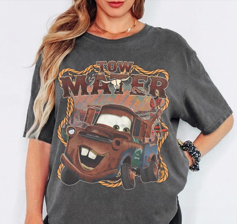 May include: Dark gray t-shirt featuring a graphic of Tow Mater from the movie Cars. The design includes the text "TOW MATER" in a western-style font, with a rope border and a bull skull.