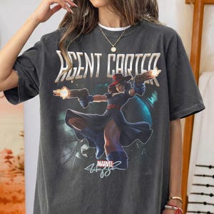 Pu&ograve; includere: T-shirt grigio scuro con la scritta "AGENT CARTER" sopra una grafica di una donna con cappello rosso e guanti blu che tiene due pistole con fiamme. Il logo Marvel e la firma sono sotto.