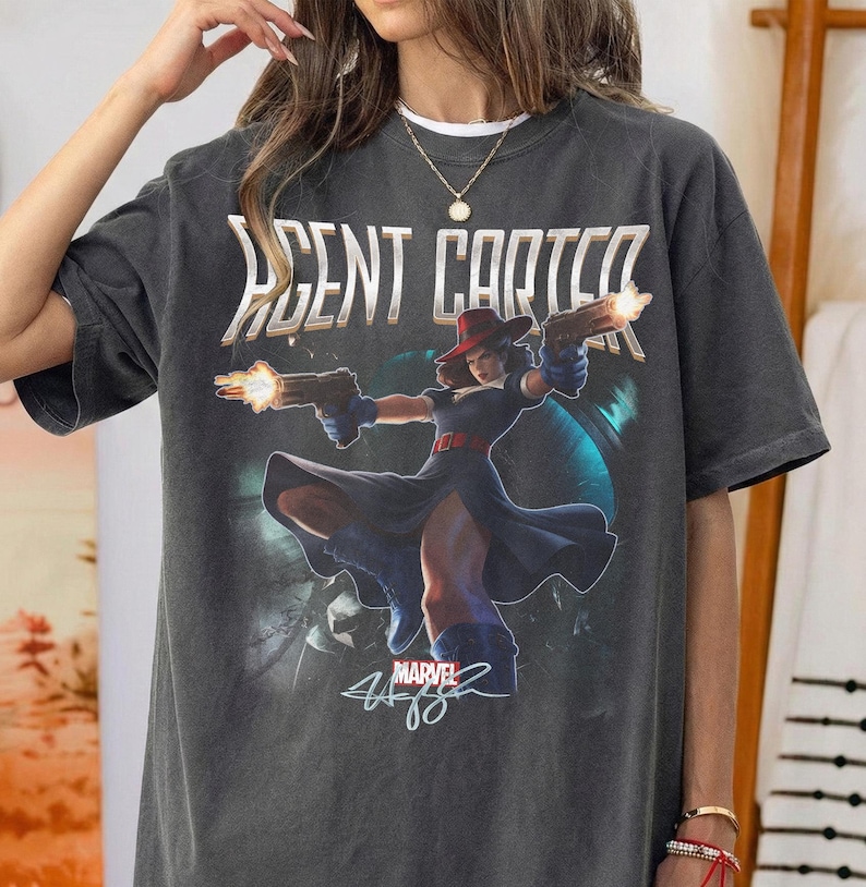Pu&ograve; includere: T-shirt grigio scuro con la scritta "AGENT CARTER" sopra una grafica di una donna con cappello rosso e guanti blu che tiene due pistole con fiamme. Il logo Marvel e la firma sono sotto.