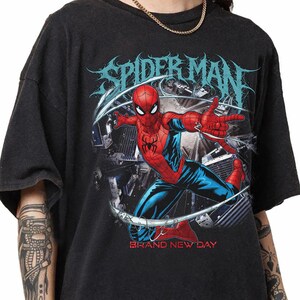 May include: Black t-shirt featuring a graphic of Spider-Man in a dynamic pose, with the words "SPIDER-MAN" in a stylized font above. The phrase "BRAND NEW DAY" is printed below the image. The superhero is depicted in his classic red and blue suit.
