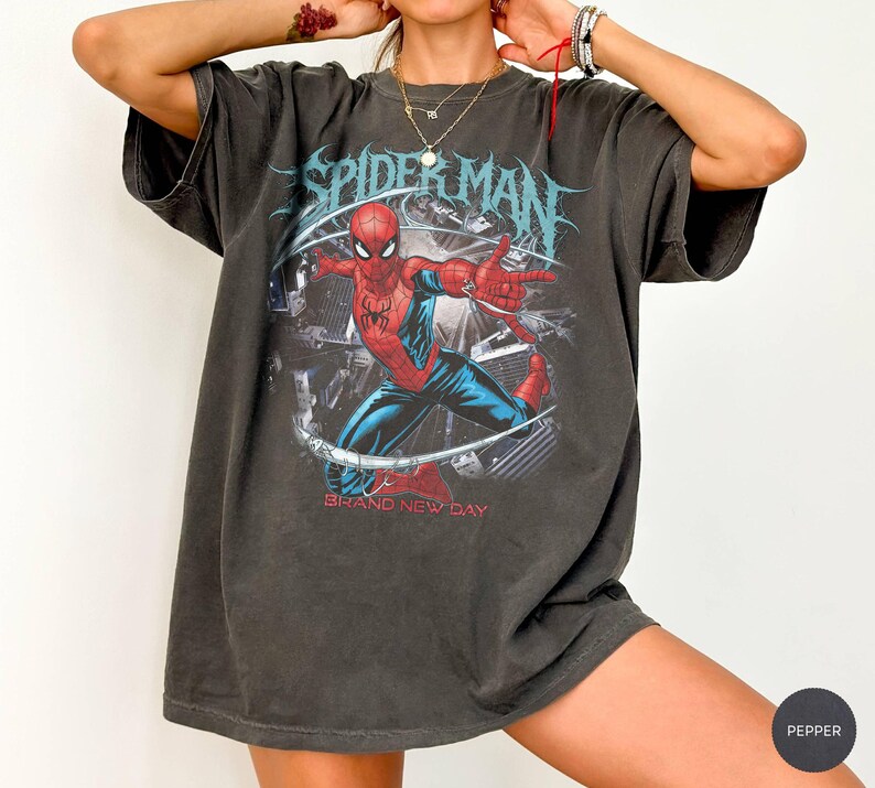 May include: Dark gray t-shirt featuring a Spider-Man graphic. The graphic shows Spider-Man in a dynamic pose with red and blue suit. The text "SPIDER-MAN" is above the image, and "BRAND NEW DAY" is below. The shirt has short sleeves and a relaxed fit.