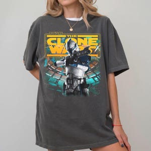 May include: A charcoal gray short-sleeve t-shirt featuring a graphic print of a Star Wars The Clone Wars character in white and blue armor holding a blaster. The design includes yellow text reading Star The Clone Wars and a teal background element.