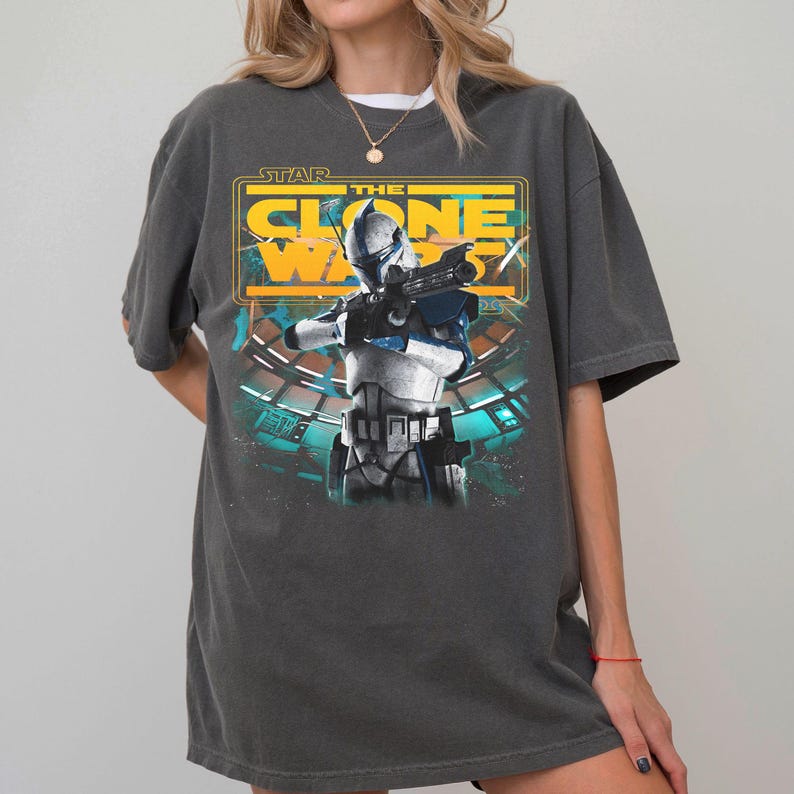 May include: A charcoal gray short-sleeve t-shirt featuring a graphic print of a Star Wars The Clone Wars character in white and blue armor holding a blaster. The design includes yellow text reading Star The Clone Wars and a teal background element.