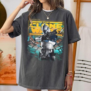 May include: A charcoal gray short-sleeve t-shirt featuring a graphic print of a Star Wars The Clone Wars character in white and blue armor holding a blaster. The design includes yellow text and a teal background with a circular structure.