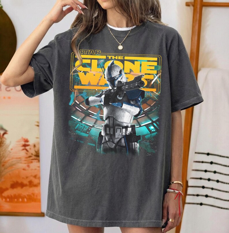 May include: A charcoal gray short-sleeve t-shirt featuring a graphic print of a Star Wars The Clone Wars character in white and blue armor holding a blaster. The design includes yellow text and a teal background with a circular structure.