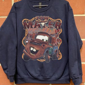 May include: Navy blue sweatshirt featuring a graphic of Tow Mater from the movie Cars. The graphic includes the text "TOW MATER" in a western-style font, with a rope border. The image of Mater is in shades of brown, red, and blue.