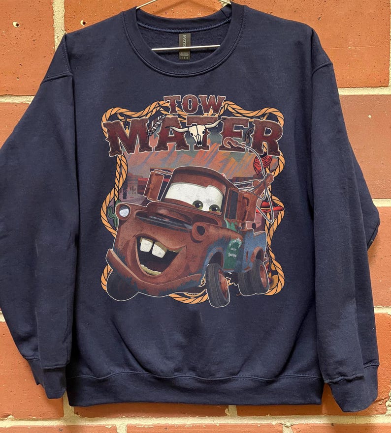 May include: Navy blue sweatshirt featuring a graphic of Tow Mater from the movie Cars. The graphic includes the text "TOW MATER" in a western-style font, with a rope border. The image of Mater is in shades of brown, red, and blue.