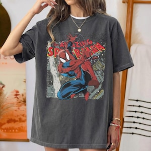 Puede incluir: Una camiseta de manga corta en color gris carb&oacute;n con un estampado gr&aacute;fico del personaje de c&oacute;mic Spider-Man en una pose din&aacute;mica. El texto "THE AMAZING SPIDER-MAN" aparece en letras rojas y amarillas sobre el personaje con un fondo urbano.