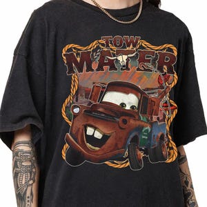 May include: Dark gray t-shirt featuring a cartoon image of Tow Mater from the movie Cars. The graphic includes the text "TOW MATER" in a western-style font, with a rope border and a desert background.