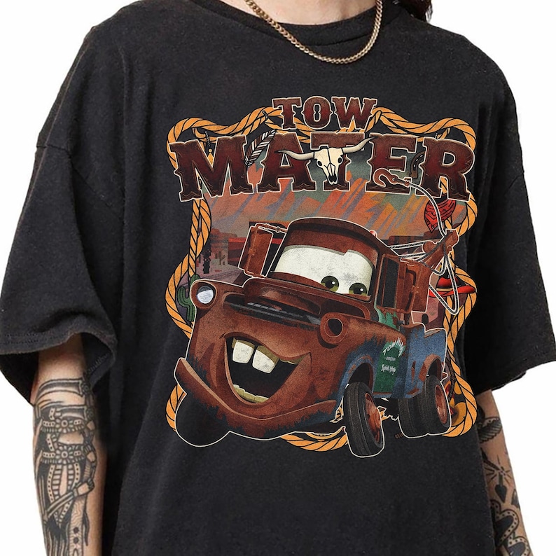 May include: Dark gray t-shirt featuring a cartoon image of Tow Mater from the movie Cars. The graphic includes the text "TOW MATER" in a western-style font, with a rope border and a desert background.