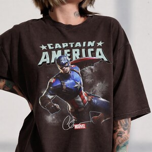 Puede incluir: Camiseta marr&oacute;n oscuro con un gr&aacute;fico de Capit&aacute;n Am&eacute;rica en acci&oacute;n. El texto "CAPTAIN AMERICA" est&aacute; impreso encima de la imagen. El logotipo y la firma de Marvel est&aacute;n en la parte inferior.