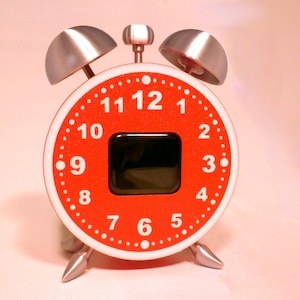 May include: A round alarm clock with a bright red face featuring white numbers from 1 to 12. The clock has a central black square display, two silver-coloured bells on top, a small silver handle, and two silver-coloured pointed legs at the base.