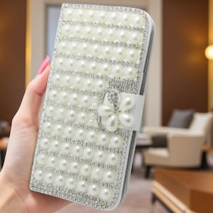 May include: A rectangular white phone case featuring a grid of round white faux pearls and rows of small clear rhinestones. A white tab closure on the side is decorated with a flower-shaped embellishment made of white pearls and clear rhinestones.