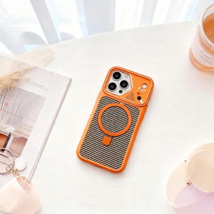 Könnte beinhalten: Eine orangefarbene Smartphone-Hülle mit einer strukturierten Rückseite im schwarz-grauen Carbonfaser-Look, einem kreisförmigen magnetischen Ladering und einem erhöhten Kameramodulschutz mit kleiner Astronautengrafik und metallischen Akzenten.