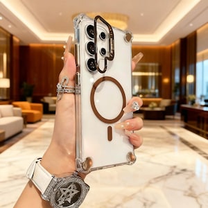 May include: A clear protective phone case featuring a brown circular magnetic ring on the back and a brown camera lens frame. The case has reinforced corners with brown accents and a transparent back panel designed for a smartphone with multiple lenses.