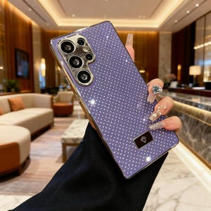 Puede incluir: Una funda de teléfono lavanda con un marco dorado y un patrón de diamantes. La funda tiene un módulo de cámara dorado y un pequeño emblema rectangular dorado con forma de diamante.