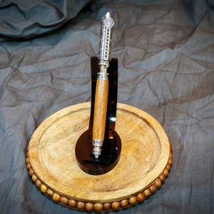 May include: A wooden pen with silver metal detailing, presented on a wooden oval tray with a beaded border. The pen has a light brown wooden barrel and is held by a black stand. The tray is approximately 20 cm in diameter.