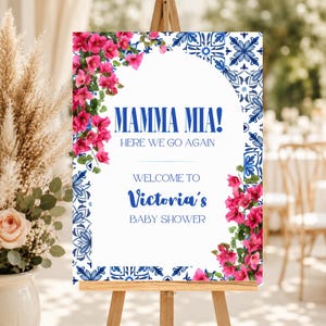 May include: A rectangular welcome sign featuring a white arched center with blue text reading MAMMA MIA! HERE WE GO AGAIN WELCOME TO Victoria's BABY SHOWER. The border displays blue and white tile patterns and clusters of vibrant pink bougainvillea.