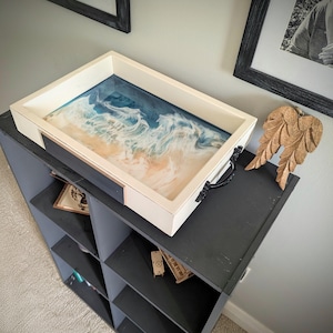 May include: A rectangular white tray with a resin ocean design. The tray has a white frame, black handles, and a resin surface depicting a beach scene with blue and tan hues. The tray is placed on a dark gray shelf.