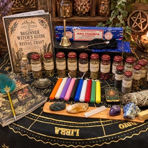 Op de afbeelding: Een verzameling rituele benodigdheden, waaronder een boek getiteld The Beginner Witch's Guide to Ritual Craft, een doos Nag Champa-wierook, een ganzenveer met een groenblauwe veer, diverse glazen potten gevuld met gedroogde kruiden, kleurrijke kaarsen, kristallen en een bundel salie op een zwart kleed met gouden dierenriemsymbolen.