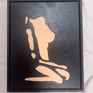 May include: A framed wall art piece featuring a black background with a peach-colored abstract silhouette of a figure in a kneeling position. The artwork is contained within a thin black frame, creating a high-contrast visual display.