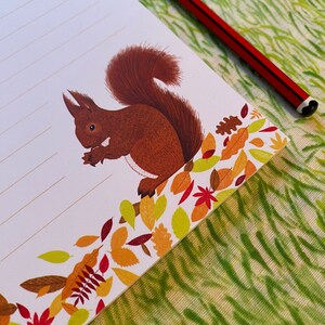 May include: A notepad with a brown squirrel illustration and a border of autumn leaves in red, orange, and yellow. A red and black pencil lies on the notepad, which is on a green patterned surface.