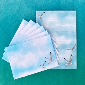 May include: A stationery set with a blue sky and cloud design, featuring watercolour artwork. The set includes envelopes and lined paper, decorated with illustrations of seagulls in flight. The envelopes are stacked in a fan shape.