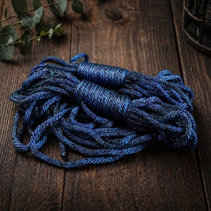 Puede incluir: Un montón de cuerda azul, probablemente para manualidades o decoración. La cuerda tiene una superficie texturizada y un color azul oscuro con reflejos azules más claros. Descansa sobre una superficie de madera oscura, con veta natural.
