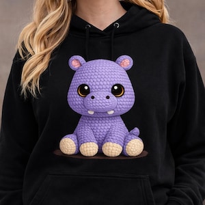 May include: Black hoodie featuring a cute, purple crocheted hippo with large, expressive eyes and small ears. The hippo has a textured appearance and is sitting on a brown surface. The hoodie has a drawstring hood.