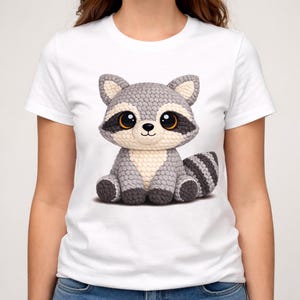 May include: A white short-sleeved t-shirt featuring a large, centered graphic of a stylized, crocheted-look raccoon. The raccoon has a grey and cream-colored body, large dark eyes with orange irises, and a striped tail, rendered in a textured, plush style.
