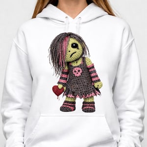 Puede incluir: Una sudadera blanca con capucha que presenta un estampado gráfico de una muñeca de ganchillo con piel verde claro, cabello oscuro con reflejos rosas y expresión triste. La muñeca viste un vestido gris oscuro con una calavera rosa y un corazón.