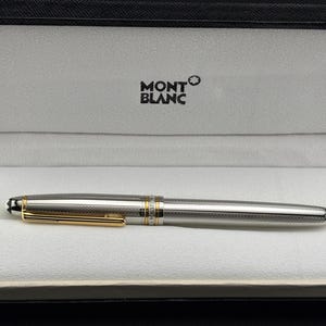 May include: A silver-toned fountain pen with a fine cross-hatched texture, featuring a gold-coloured clip and accents. The pen is displayed inside a black presentation case with a white interior, which has the Montblanc logo printed in black.