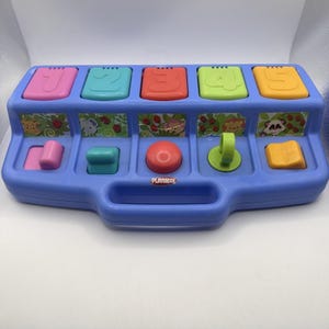 May include: A blue Playskool pop-up toy with five colorful buttons numbered 1-5. Each button is a different color: pink, teal, red, green, and yellow. Below the buttons are animal-themed pop-up shapes. The toy has a handle and the Playskool logo.