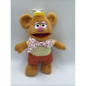 Può includere: Un peluche a forma di personaggio con grandi orecchie, naso rosa e occhi ricamati. Il personaggio indossa un cappello giallo, un papillon a pois e pantaloncini arancioni. Il giocattolo ha una camicia giallo chiaro e pantaloncini arancioni. La bocca del personaggio è aperta.